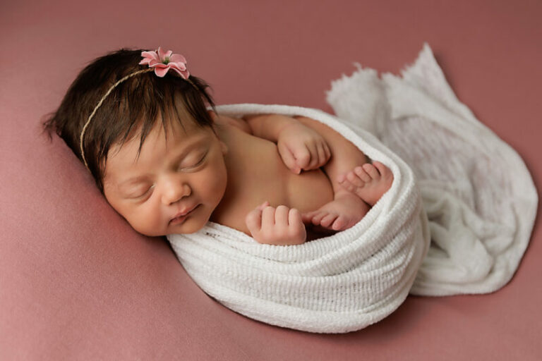 newborn girl on pink backdrop with pink flower and white wrap