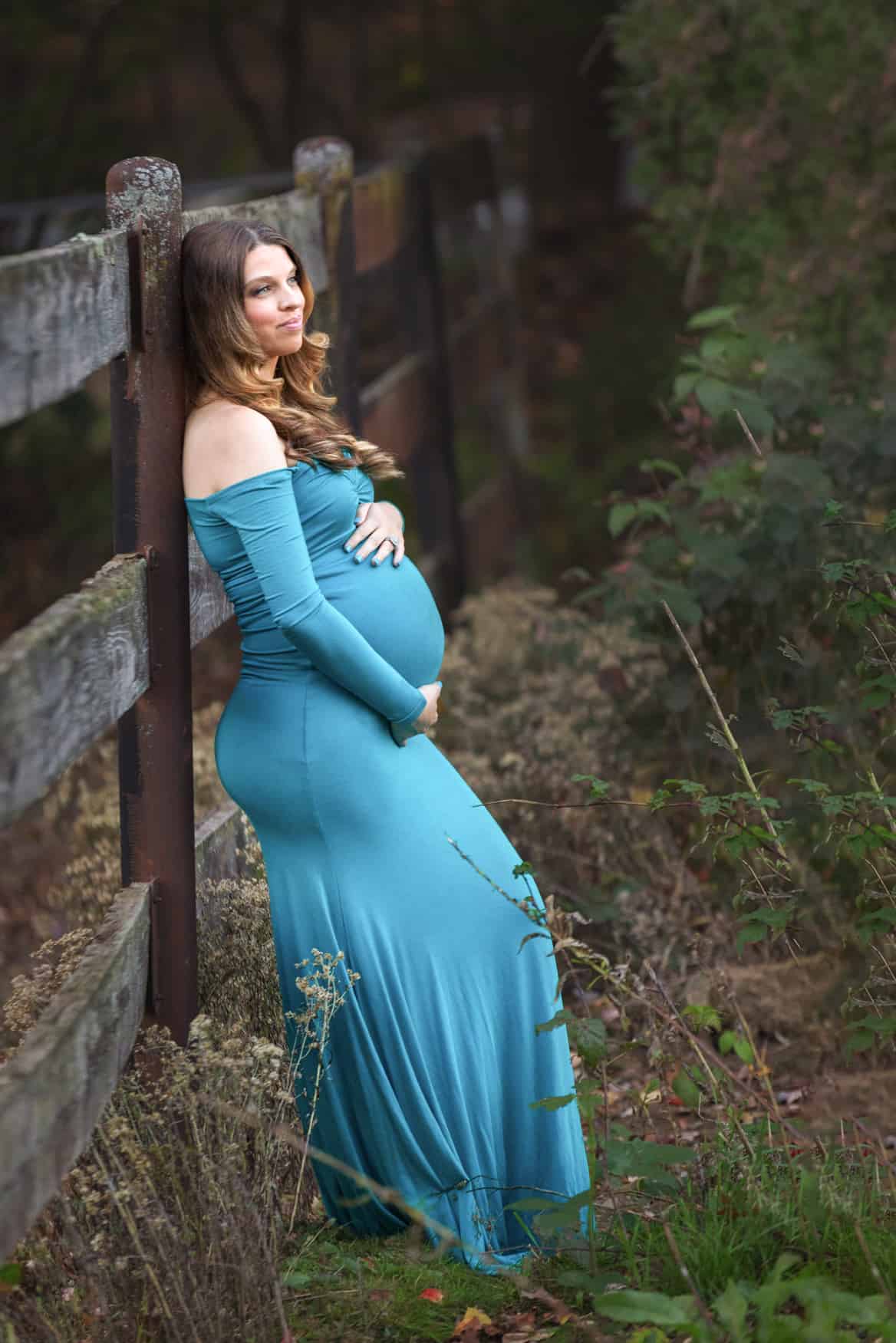 Windham NH Maternity in blue dress at horse farm leaning up against fence
