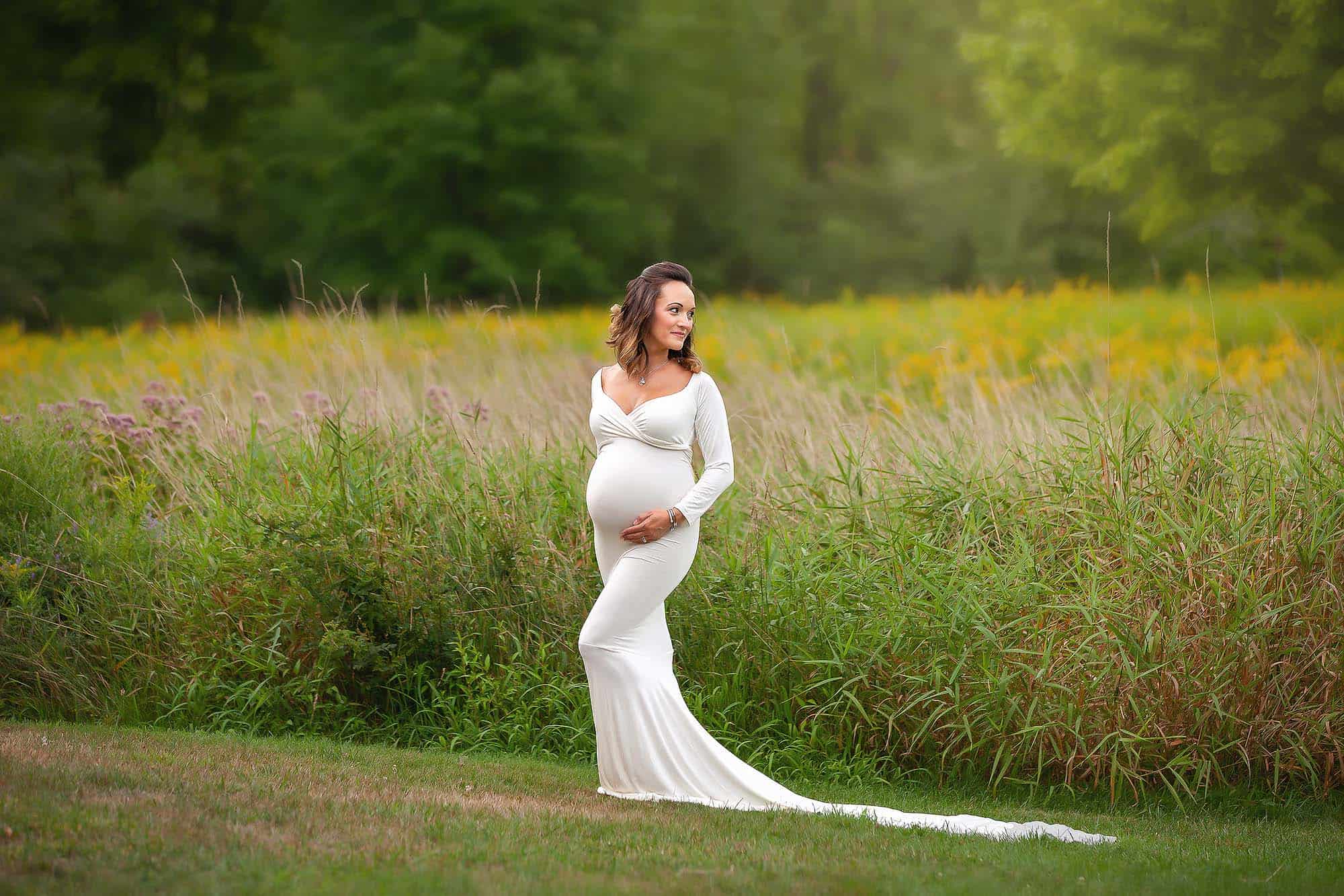 Derry NH Maternity Portrait. Pregnant woman standing in open field of tall green grass wearing a white dress.