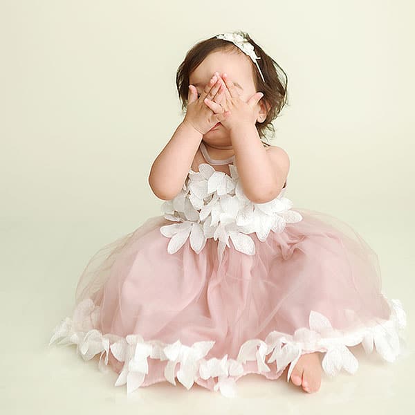 One Year Portrait of baby girl sitting on ivory backdrop in pink floral dress with hands over eyes playing peekaboo.