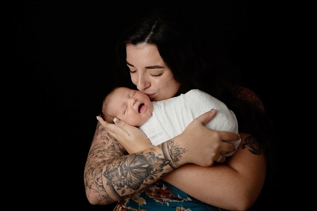 Derry NH Newborn Portrait of mother holding baby in an ivory wrap kissing him. Mother has tattoos on her arms. Black background.