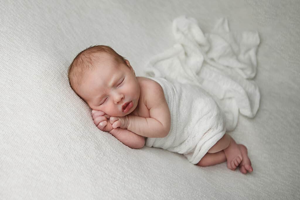Portrait of a newborn baby boy sleeping on his side with hands up under his chin wearing an ivory wrap.