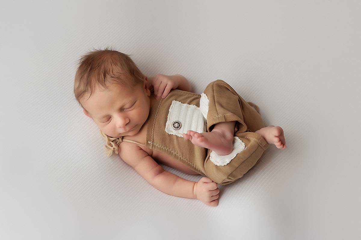 Derry NH Newborn Portrait. Baby Boy laying on ivory backdrop wearing a tan romper. Baby is sleeping on his back in Huck Finn pose.