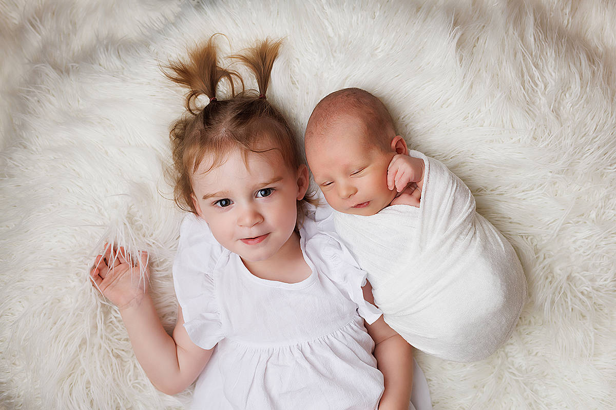 Newborn in ivory wrap laying on a fur throw blanket with his 2 year old sister snuggled up next to him for a newborn portrait. Baby Safety is so important when working with young siblings.