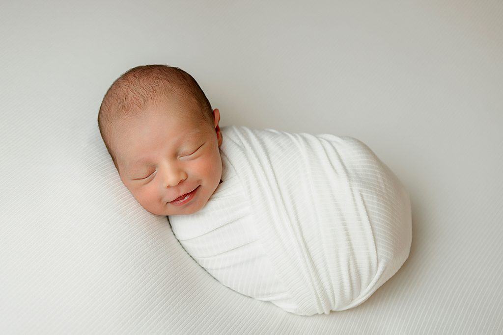 Newborn wrapped in ivory wrap on ivory backdrop. Smiling