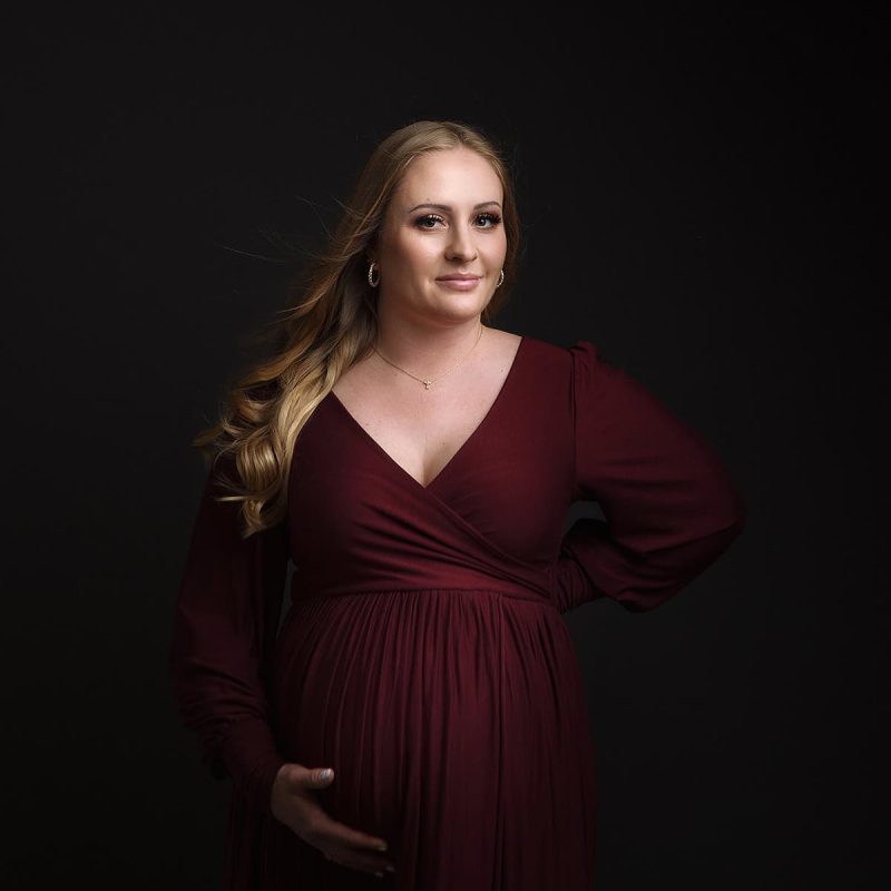 Windham NH Maternity Portrait - Mother to be looking at the camera with her hand on her belly in a maroon couture maternity gown.