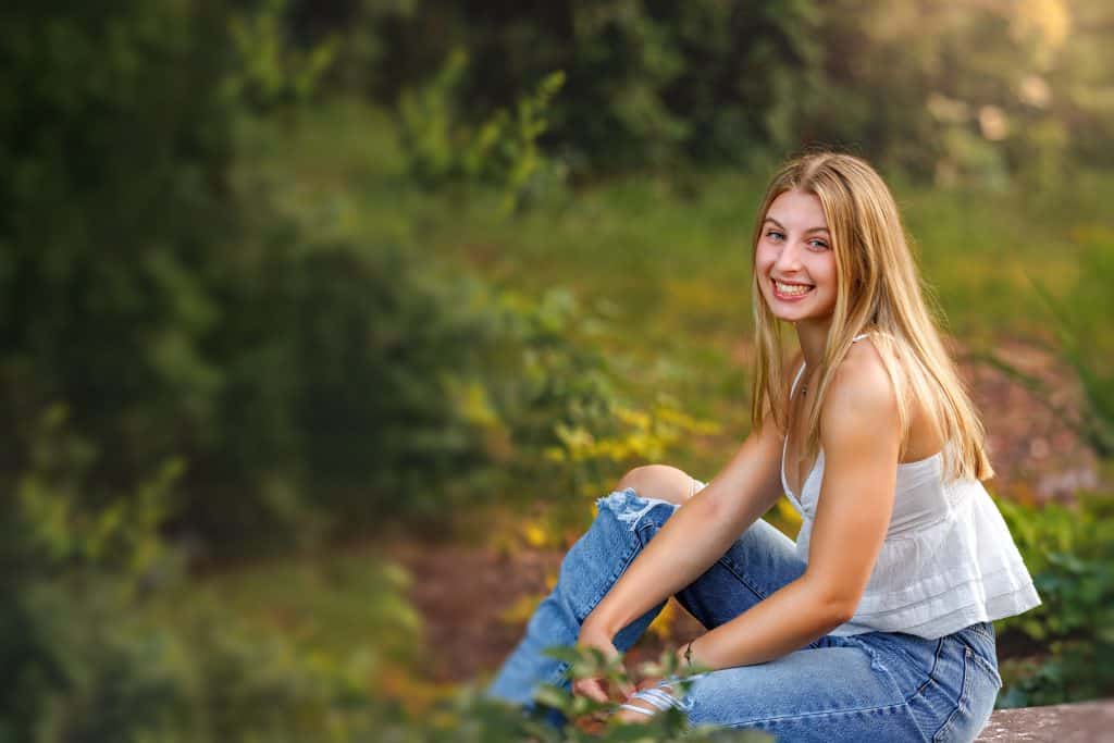 Teenage Senior Portrait of girl sitting on bench wearing blue jeans with ripped knees and a white tank top blouse.