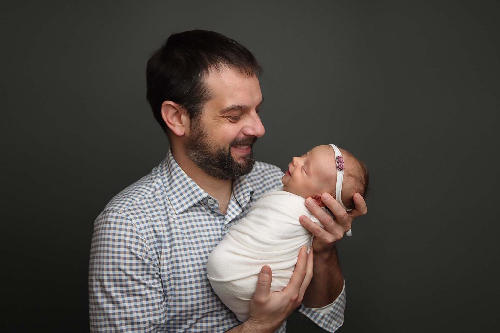 Salem NH Newborn Portrait Session of baby girl swaddled in a white wrap being held by her father. Both father and baby are smiling.