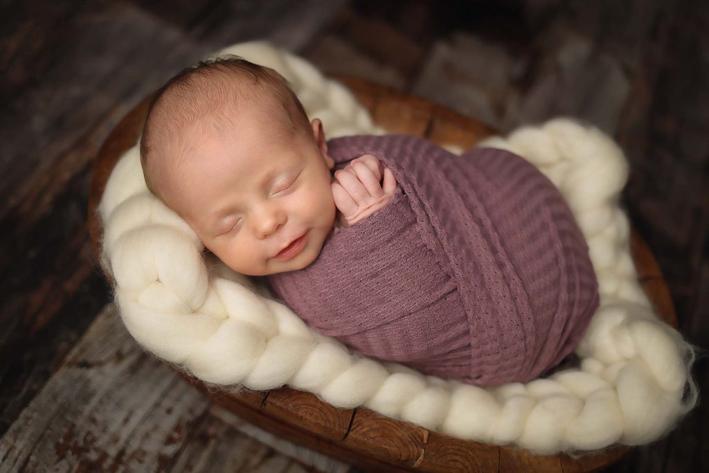 Salem NH Newborn Portrait of baby girl swaddled in a purple wrap. Laying in a wooden bowl.