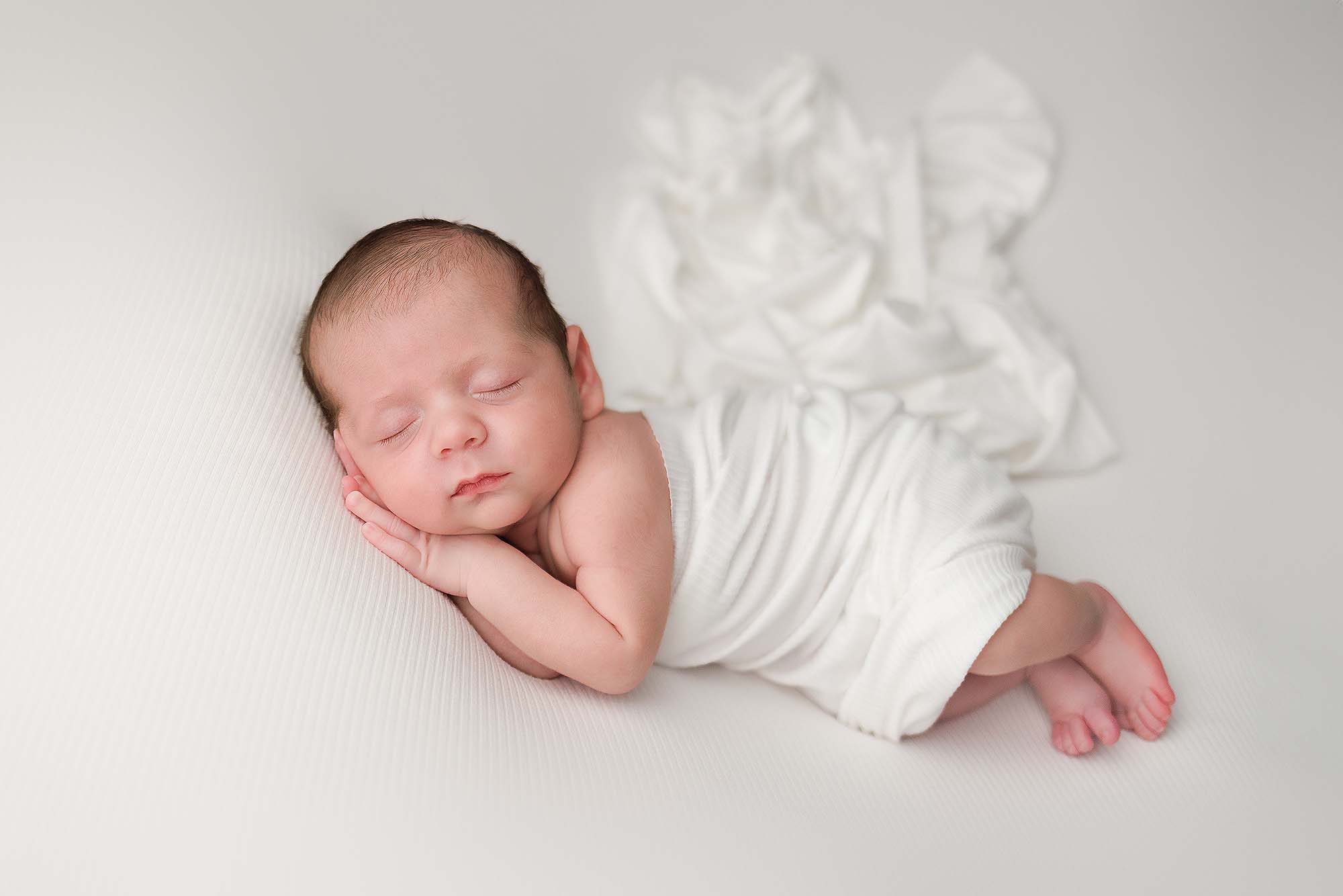 Newborn wrapped in a ivory wrap in a side pose with hands tucked under cheeks.