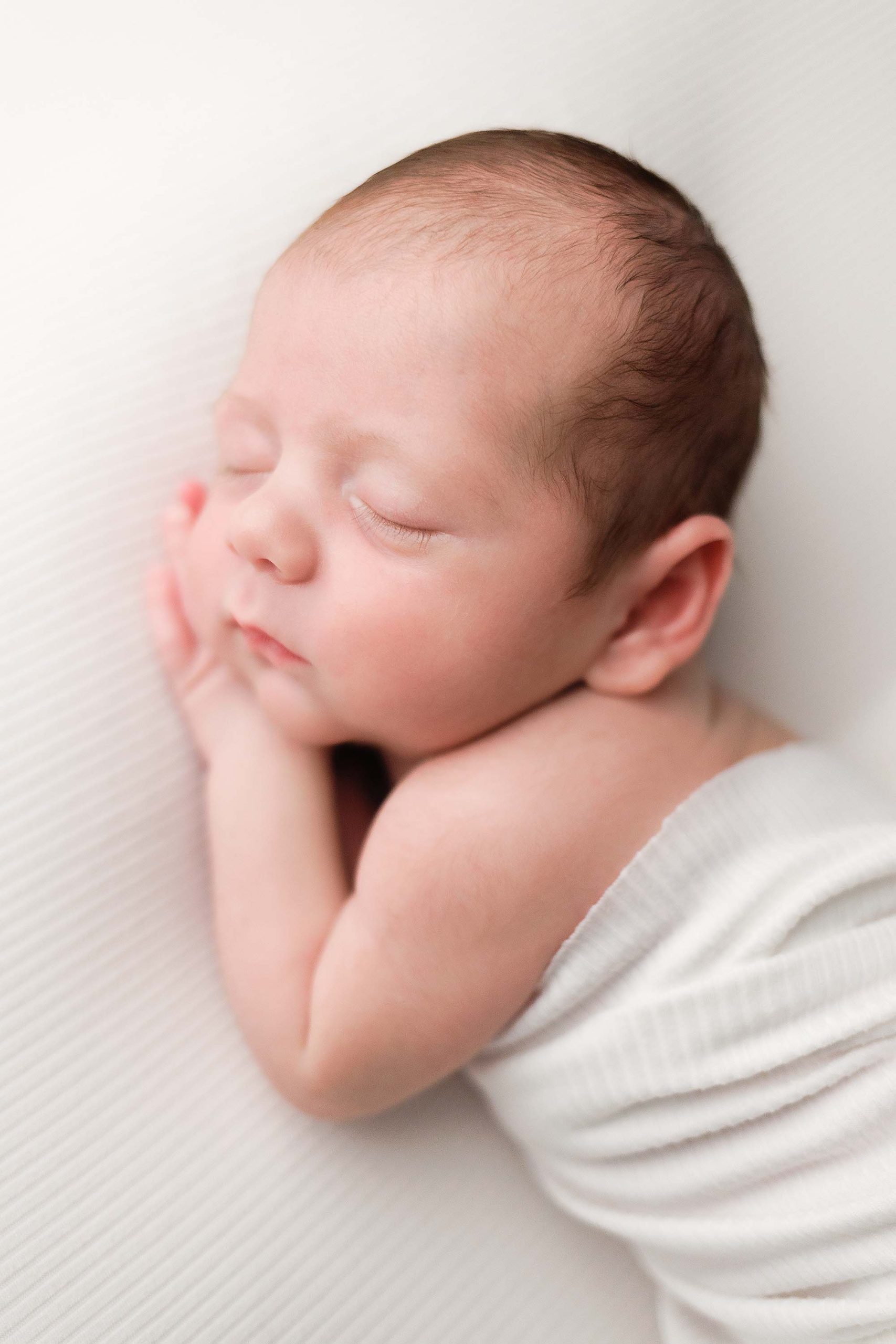 Boston Newborn Portrait of a baby boy on ivory background in an ivory wrap sleeping with his hands tucked under the side of his face.