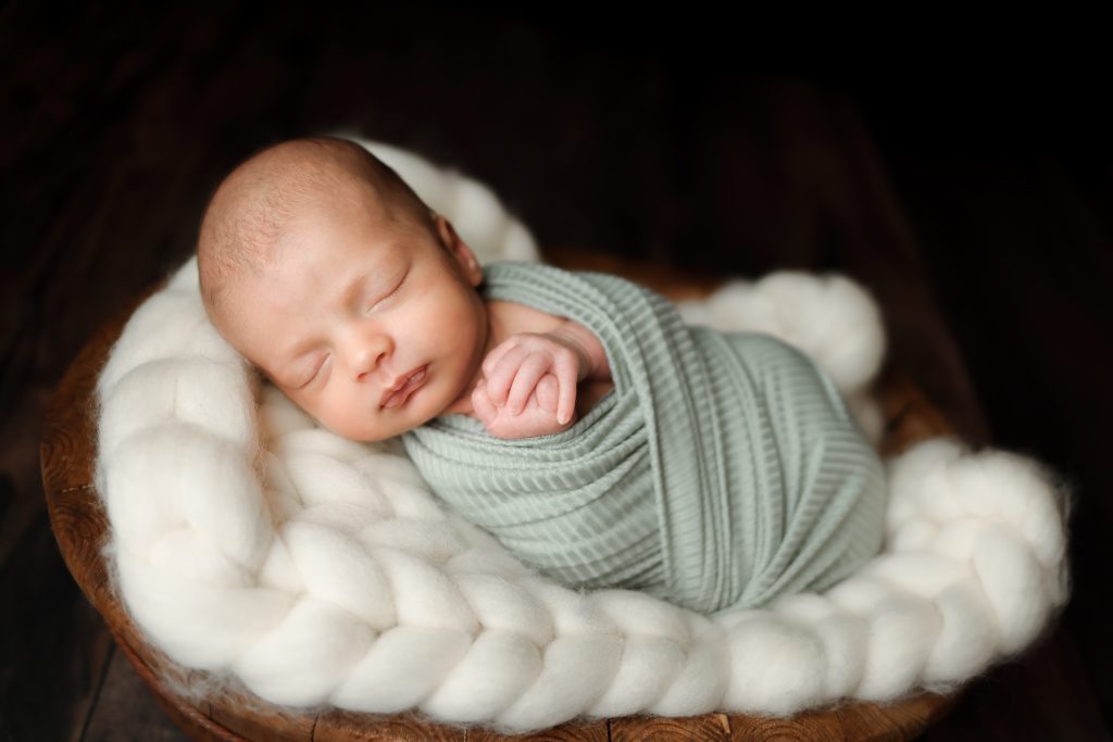 Newborn Portrait of baby boy swaddled in sage wrap laying ontop of an ivory large knit bed in a wooden basket.