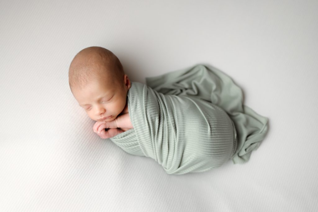 Swaddled newborn in a sage green wrap. Portrait taken from above.