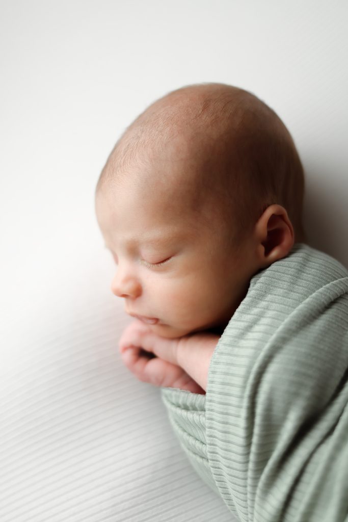 Boston Newborn Portrait of baby boy laying on side swaddled in a sage green wrap with a soft light illuminating his profile.