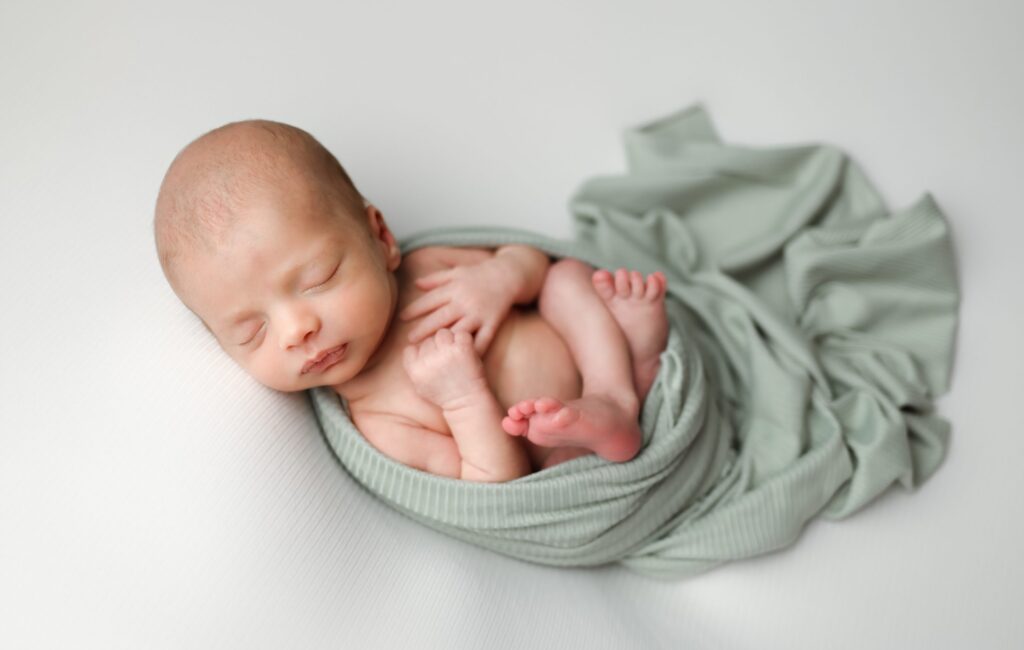 Boston Newborn portrait of baby boy swaddled in a sage green wrap laying on his back with hands and feet showing in a womb pose.