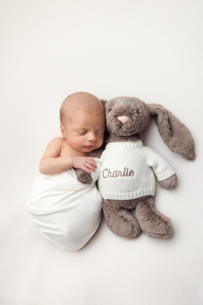 Boston Newborn portrait of baby boy laying on his back snuggled up to a soft stuffed bunny toy wearing a sweater with baby's name on it.