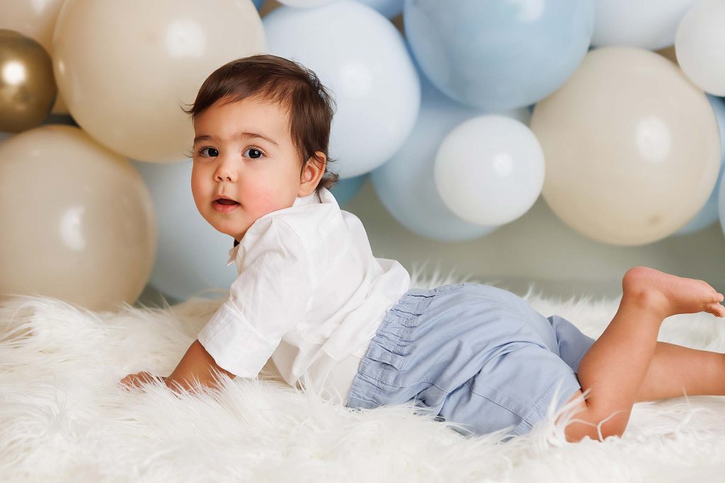 One Year cake smash session of little boy laying on his tummy on a white fur throw.