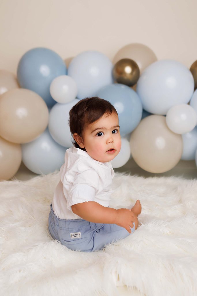 One year portrait of baby boy sitting and turning to look at the camera. Sitting on a white fur throw with balloons in the background.