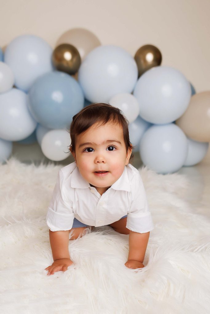 One Year Cake smash in Southern New Hampshire of little boy crawling on a white fur throw with balloons in the background.