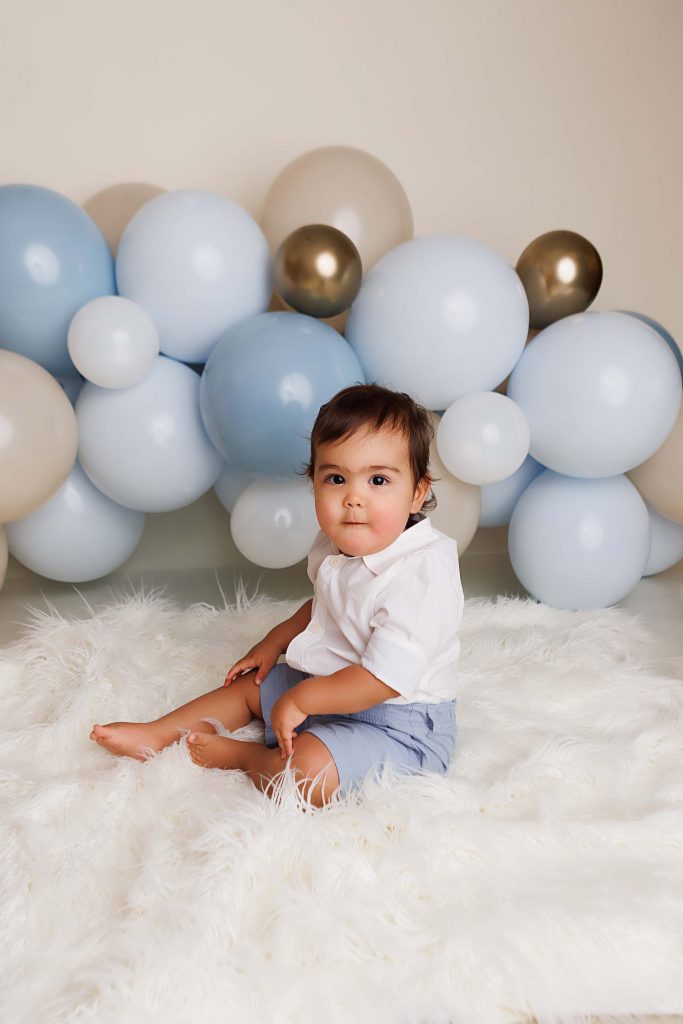 Cake Smash session near me of one year old sitting on fur throw with balloon garland in background.