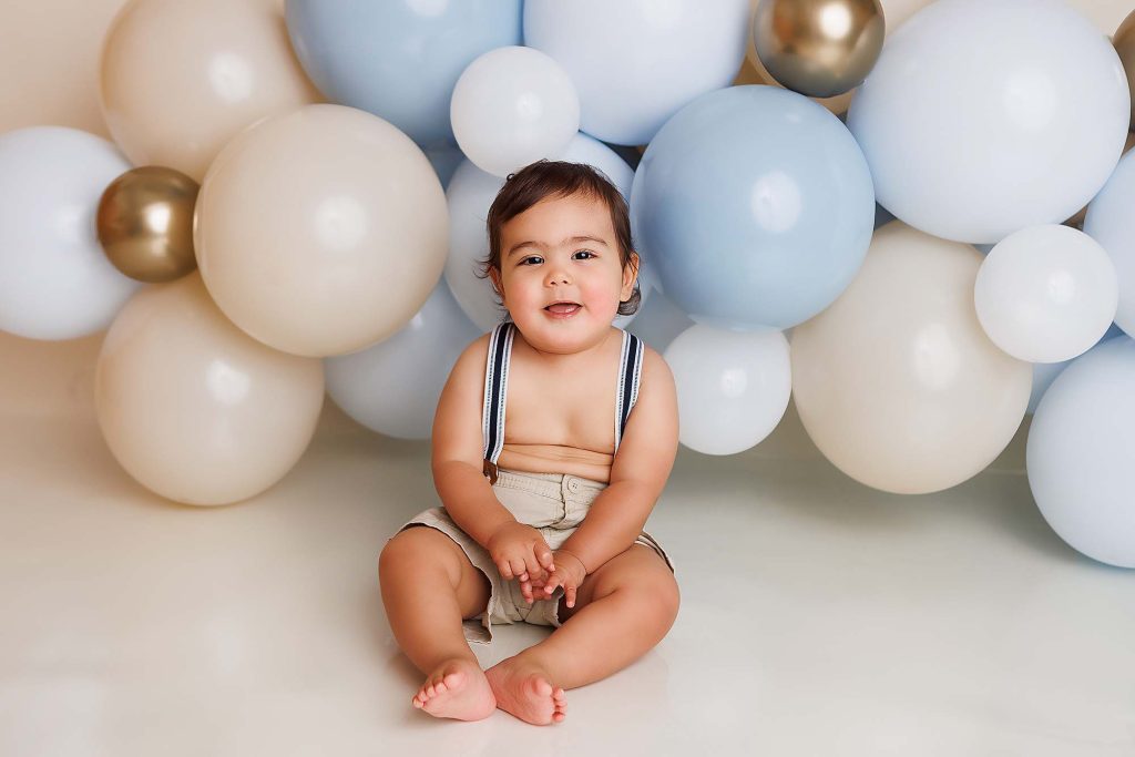 One year old sitting in front of a balloon garland wearing suspenders.