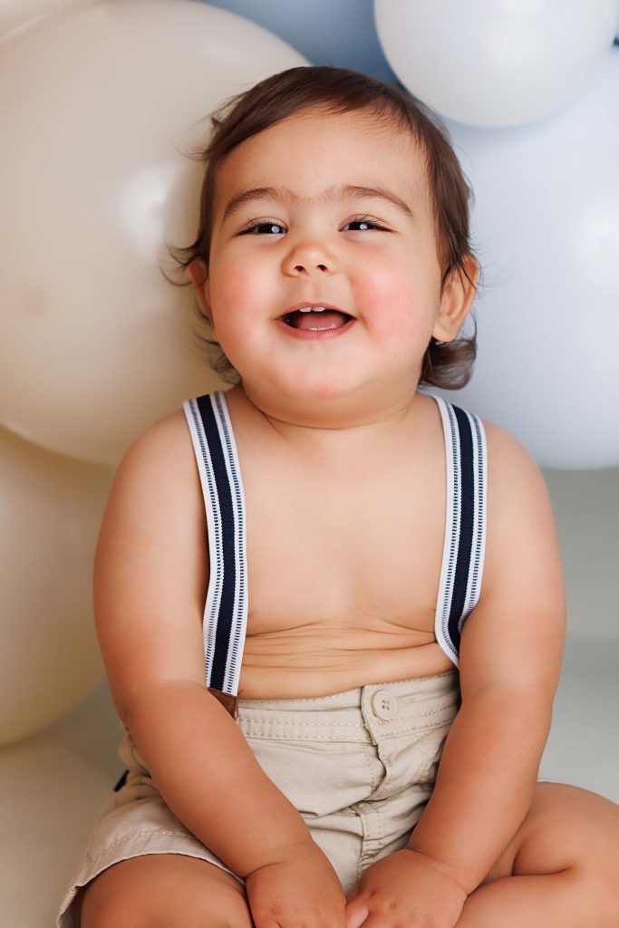 One year old laughing at the camera with balloons in the background.