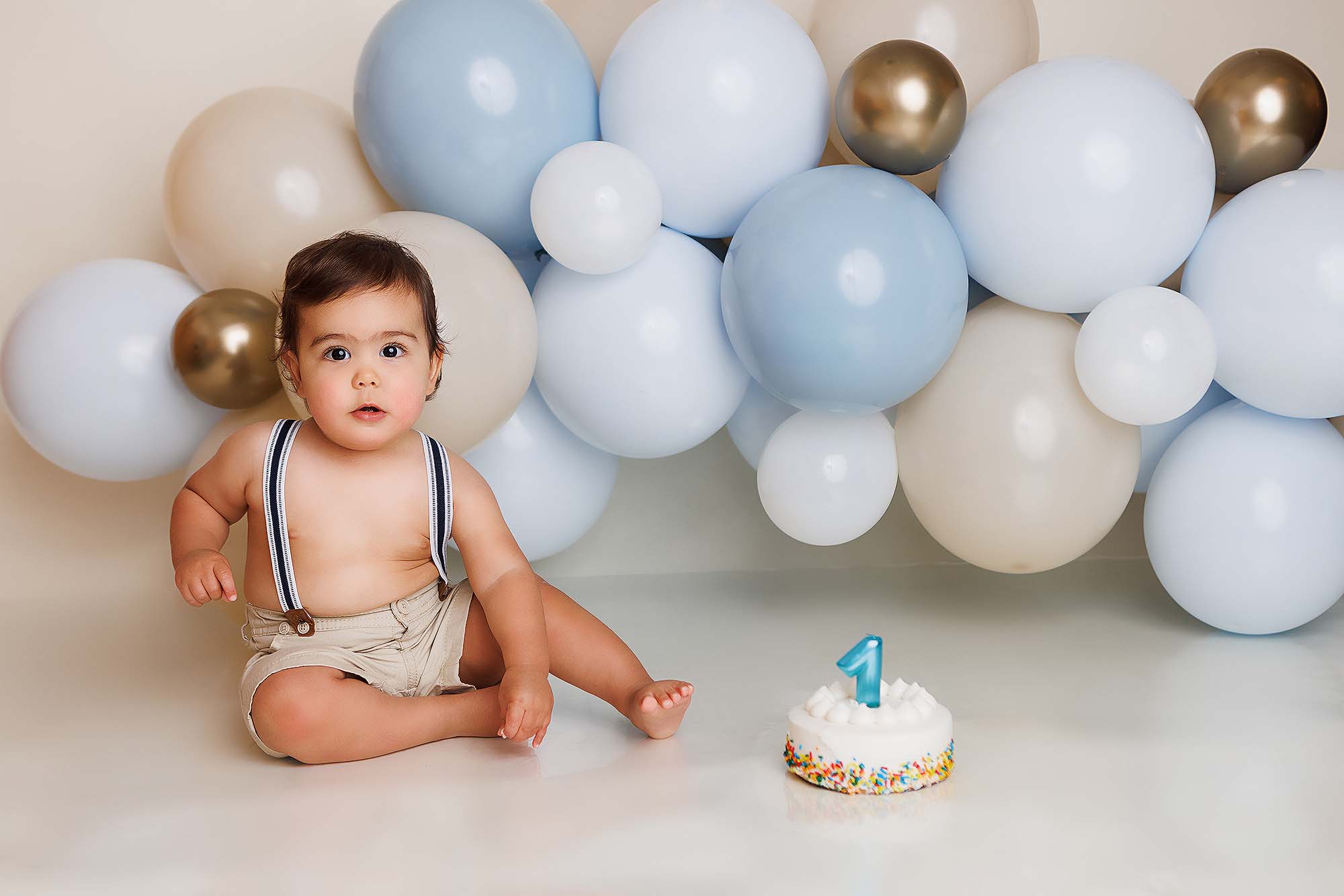 Cake smash session. Little boy sitting next a cake with balloons in the background.