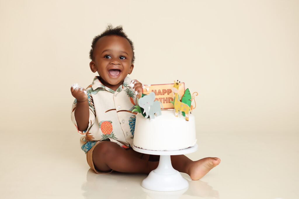 One Year old smiling at camera while sitting behind a white frosted cake for his cake smash portrait session.