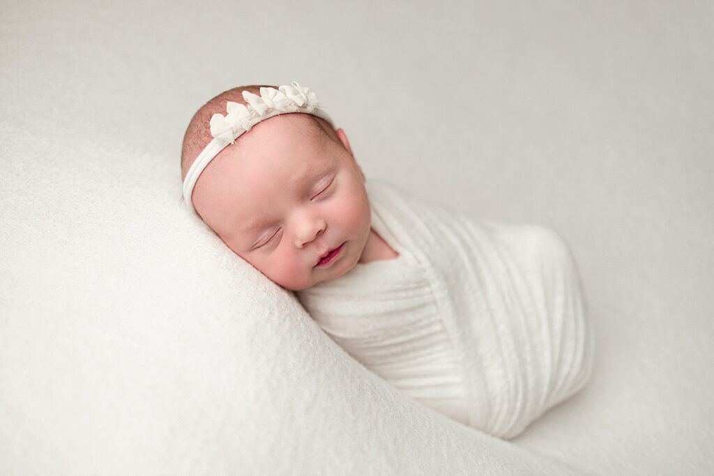 Andover Newborn Portrait of newborn baby girl swaddled in an ivory wrap wearing an ivory headband with tiny bows.