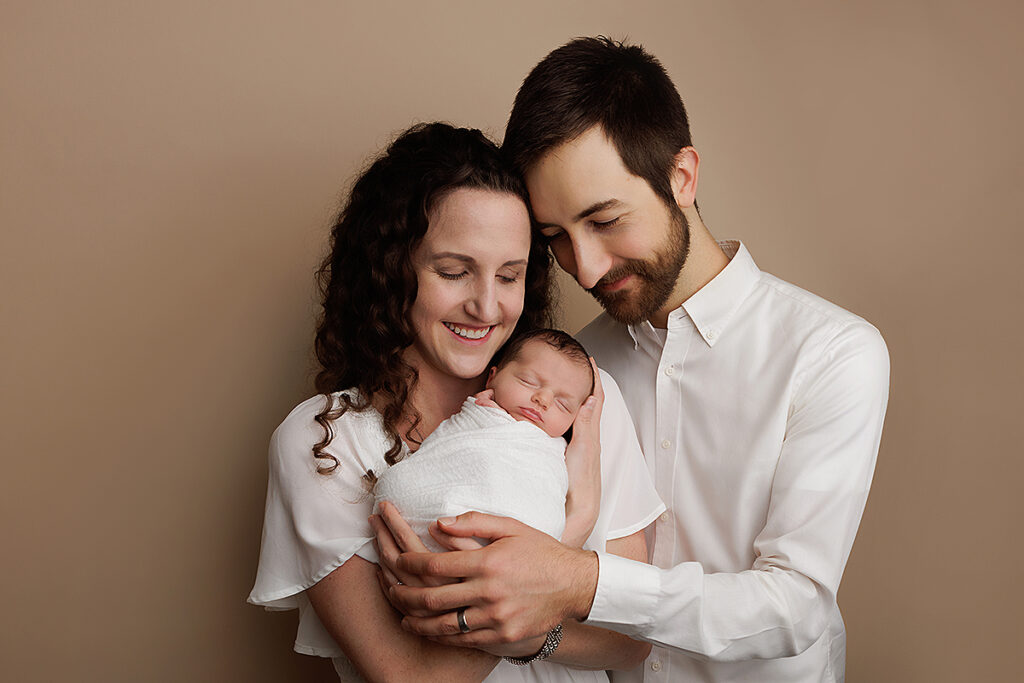 Andover Newborn Portrait of baby girl swaddled in the arms of her mother and father. Parents are looking down at her smiling.