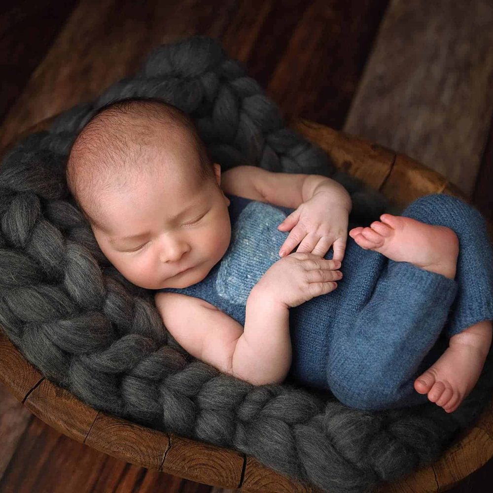 Boston Newborn Portrait of a baby boy in a blue romper. The baby is in a wooden bowl on a gray knit blanket.