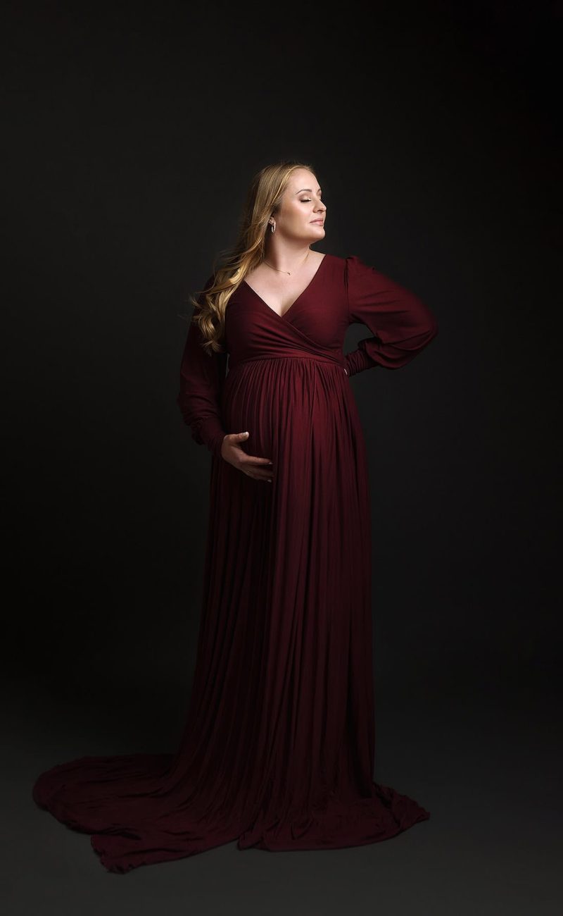 Boston Maternity Portrait of Mother to be Looking away from camera with head pointed up at light. Hand on belly wearing maroon couture maternity gown.