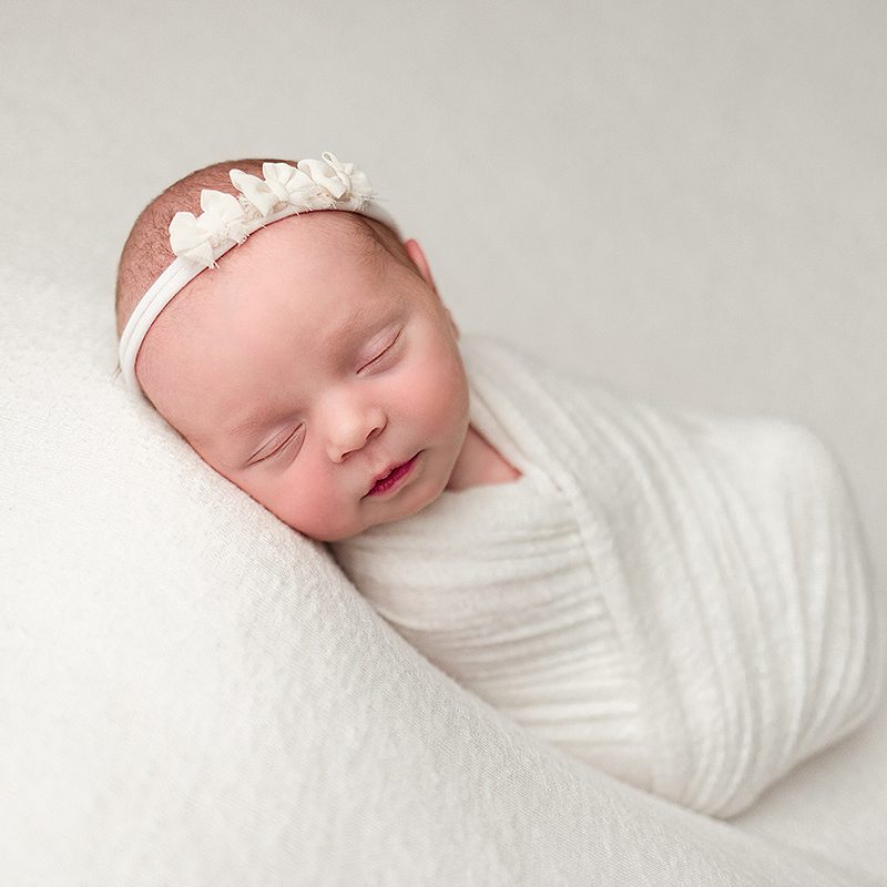 Andover Newborn Portrait of newborn baby girl swaddled in an ivory wrap wearing an ivory headband with tiny bows.