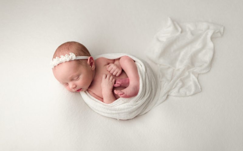 Boston Newborn Portrait of newborn baby girl wrapped in ivory wrap on her back in the a womb like pose wearing an ivory headband.