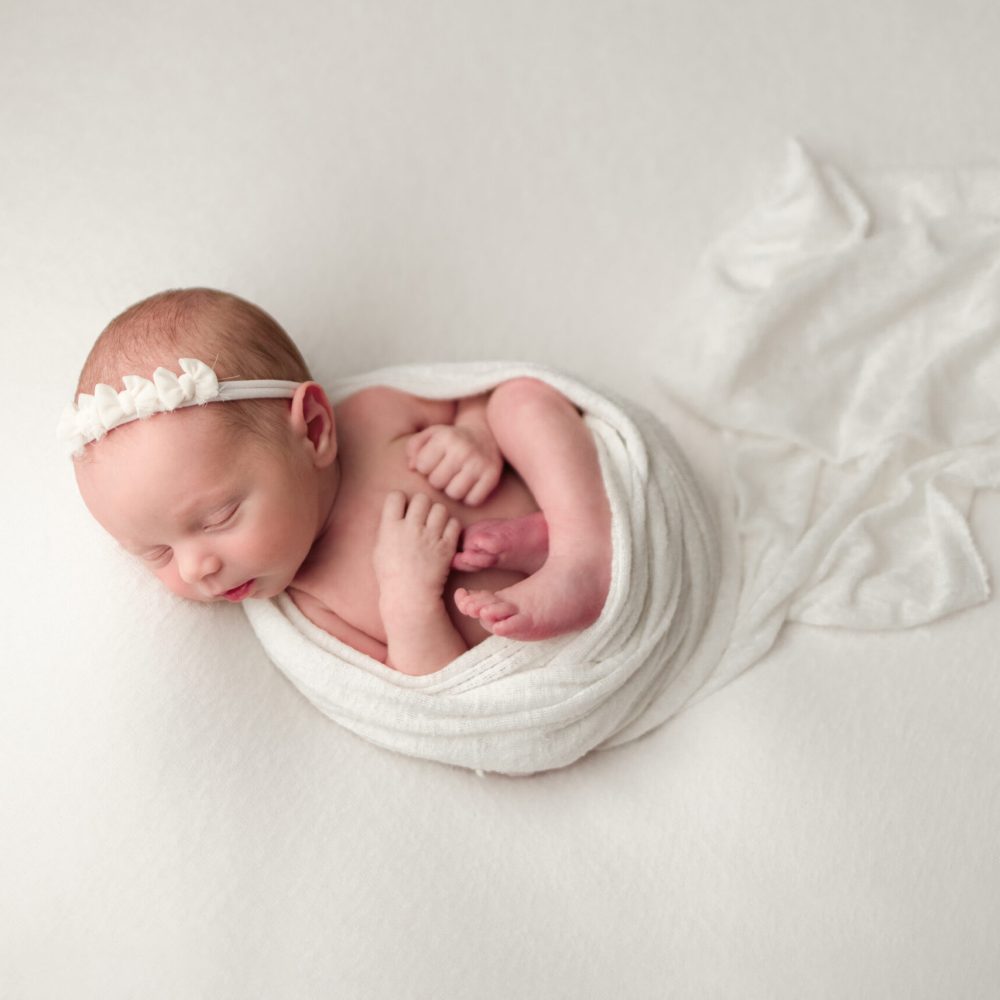 Boston Newborn Portrait of newborn baby girl wrapped in ivory wrap on her back in the a womb like pose wearing an ivory headband.
