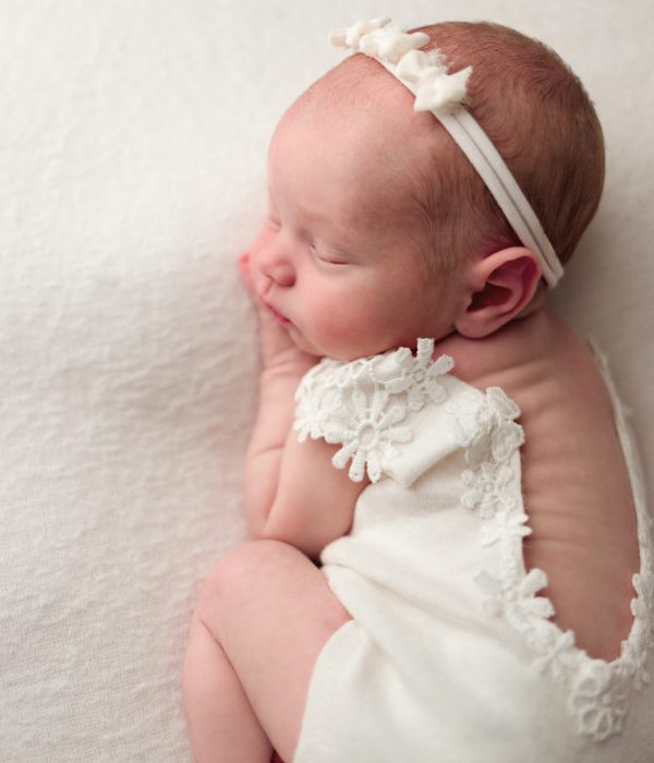 Newborn portrait near me of baby girl wearing an ivory romper and an ivory headband, sleeping all curled up.