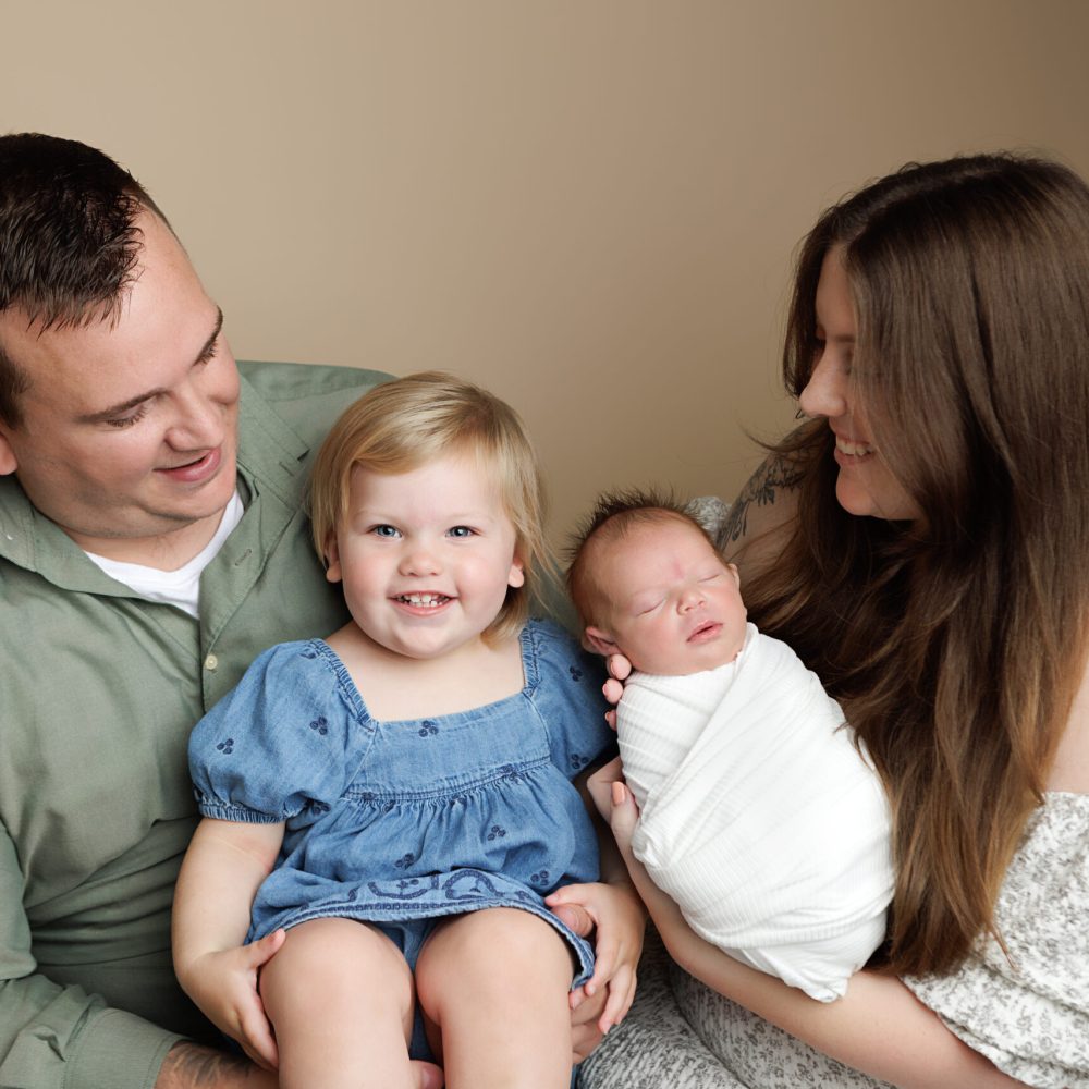 Family Portrait during a newborn session. Boston Newborn Portrait of toddler smiling at camera while mom and dad are holding newborn sister.