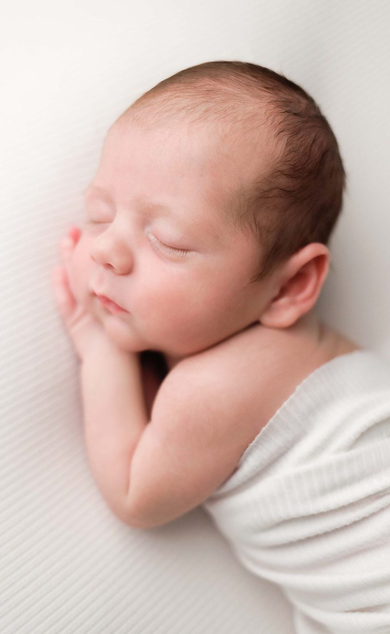 Boston Newborn Portrait of a baby boy on ivory background in an ivory wrap sleeping with his hands tucked under the side of his face.