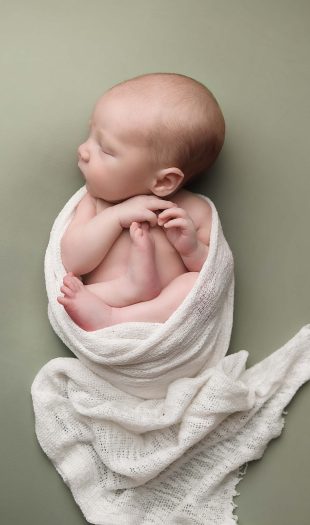 NH Newborn Portrait of baby boy on sage green background in womb pose with ivory wrap surrounding him.