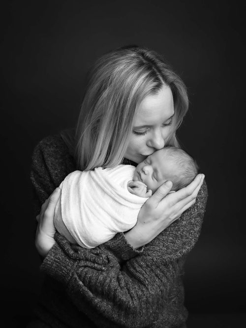 Black & White Newborn Portrait of mother holding newborn baby girl, kissing the side of her head. Baby is wrapped in a white swaddle.