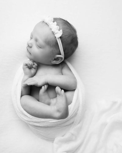 B&W Newborn Portrait of baby girl laying on her back in a womb position wrapped in a white wrap and wearing a headband with small bows.