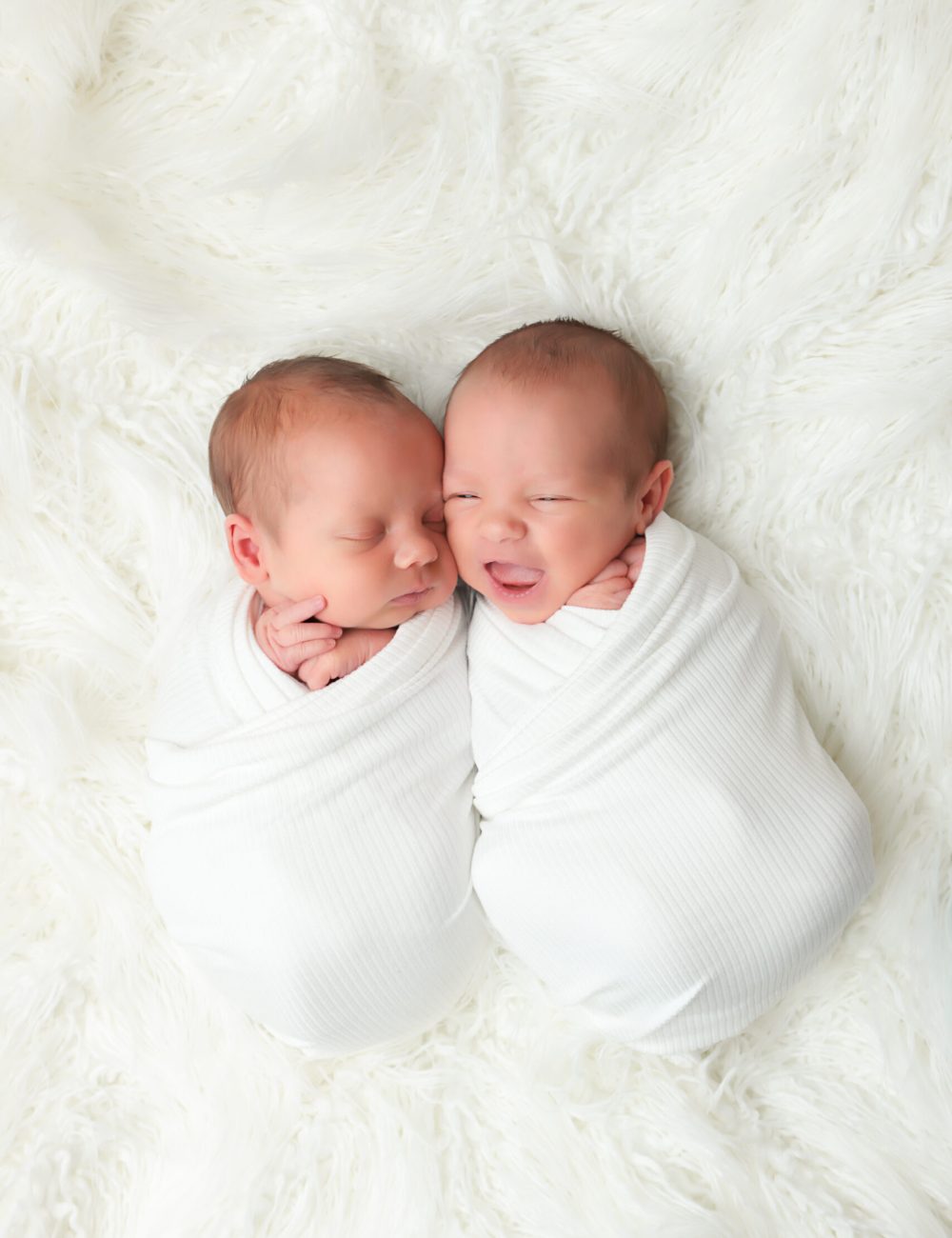 NH Twins wrapped in ivory swaddle laying next to each other on a fur throw blanket. One twin is smiling.