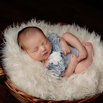 Newborn baby girl from Boston MA posing in brown basket filled with ivory fur. Baby is wearing a blue lace romper with ivory flowers.