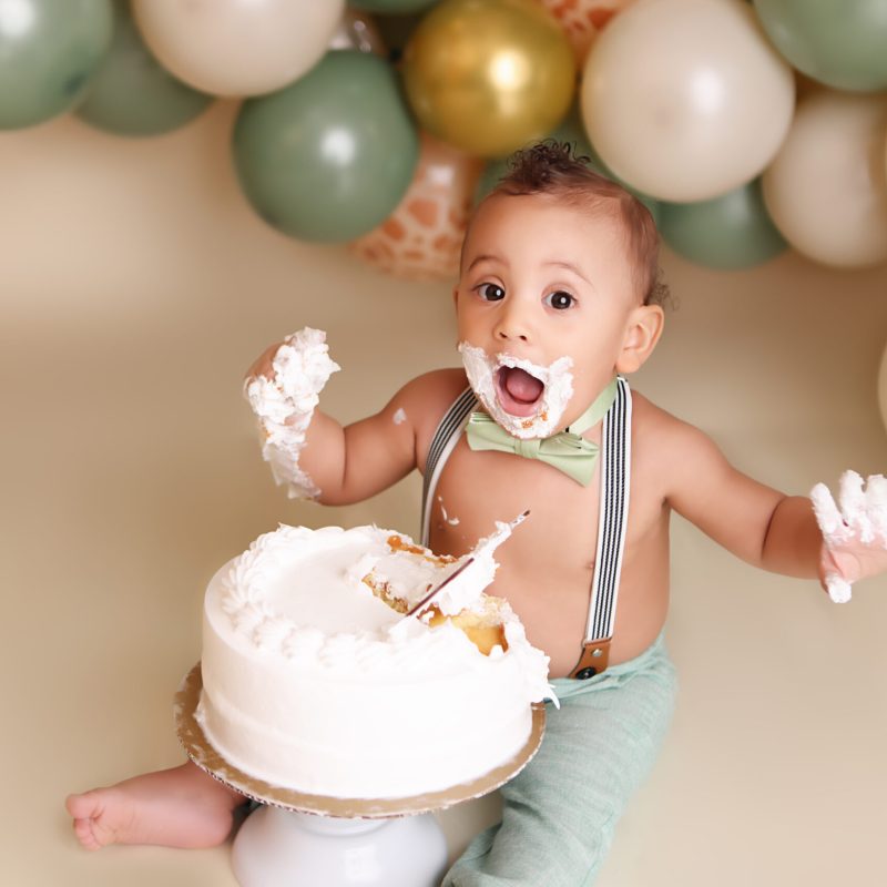 Boston One year cake smash session of little boy wearing sage green pants and suspenders while eating a white frosted cake with his hands.