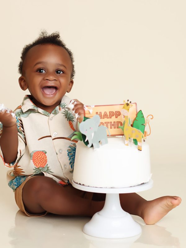 One Year old smiling at camera while sitting behind a white frosted cake for his cake smash portrait session.