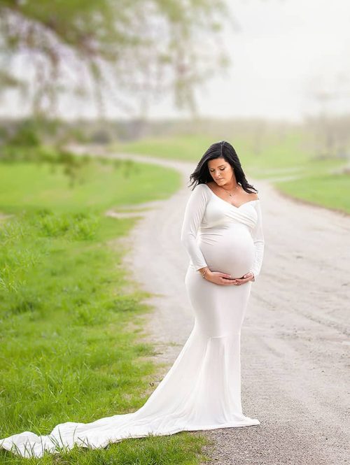 Londonderry NH Maternity Portrait. Woman Standing on edge of dirt road in white fitted long dress looking at stomach.